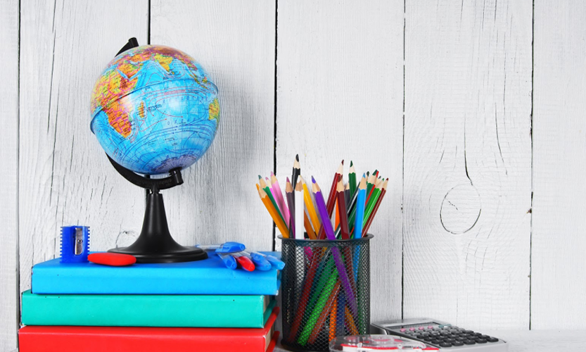 globe sitting on a desk on top of a stack of books and next to a tin filled the colored pencils